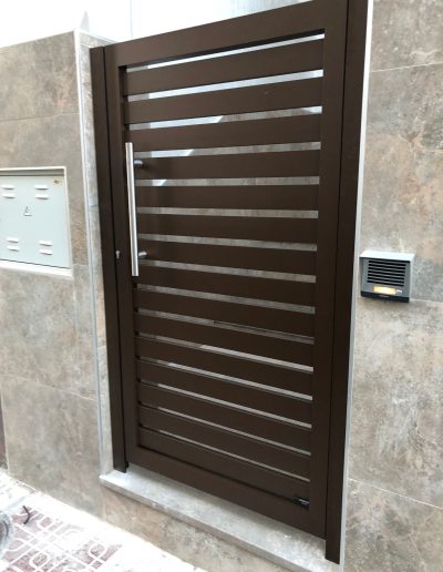 Puerta peatonal exterior de lamas metálicas con acabado texturizado